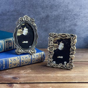May include: Two antique bronze picture frames with oval and rectangular shapes, each adorned with rose and leaf detailing. The frames hold vintage black and white portraits. One frame rests on a blue book with gold lettering.