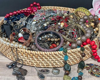 May include: A round woven wicker basket filled with a large collection of assorted jewelry, including strands of red, blue, and white pearls, metallic chains, beaded necklaces, a purple rhinestone bangle, and various pendants and brooches.