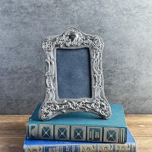 May include: A vertical rectangular picture frame featuring an ornate, polished silver-toned metal border with intricate scrollwork and floral relief patterns, set against a dark gray velvet backing with a central dark blue display area.