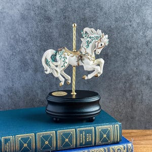 May include: A white carousel horse figurine with gold accents and green leaf detailing, mounted on a gold-colored pole and a round black base. The horse features a flowing mane and tail, with intricate decorative elements on its saddle and harness.