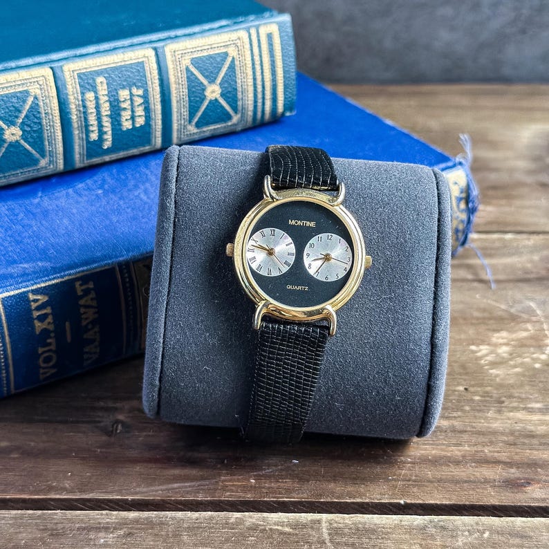 May include: A Montine quartz wristwatch featuring a gold-tone round case and a black textured strap. The black watch face displays two circular sub-dials, one with Roman numerals and one with Arabic numerals, positioned side by side on the dial.