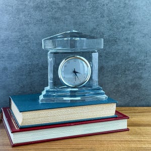 May include: A clear glass mantel clock with a rectangular base and a pediment top. The clock face is circular with a silver-toned rim, black numerals, and black hands. The clock is positioned on top of two stacked hardcover books.