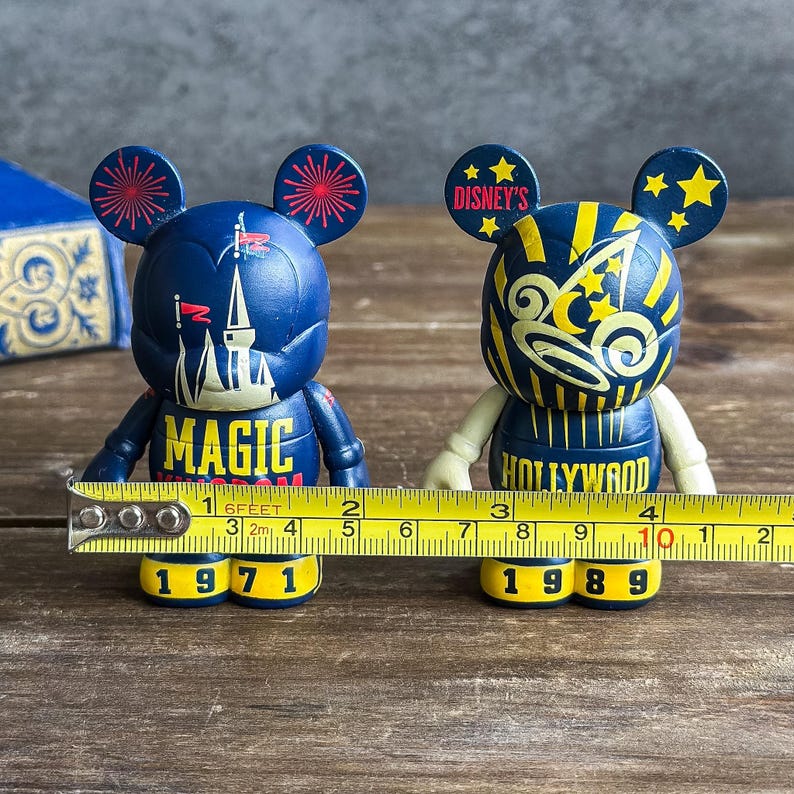 May include: Two Mickey Mouse figurines. One is navy blue with "MAGIC" and a castle design, the other is navy blue with "HOLLYWOOD". Both have yellow accents and the years 1971 and 1989 on the base. The figurines are approximately 10 cm tall.