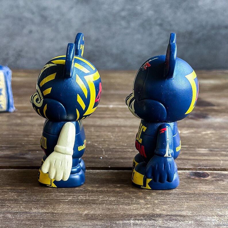 May include: Two navy blue collectible figurines with yellow and red detailing. The figurines feature a stylized bear design with a unique pattern on the head and body. One figurine has a white glove design on the arm.