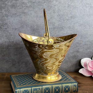 May include: A vintage brass flower bucket with a handle. The bucket has a flared shape and is decorated with an etched pattern. It sits on a blue book with gold lettering. The bucket is approximately 8 inches tall.