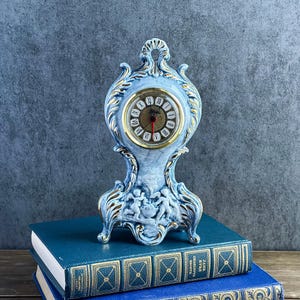 May include: A light blue ceramic mantel clock with gold trim and ornate scrollwork. The clock face features Roman numerals and a red second hand. The base displays a relief sculpture of two cherubs. The clock rests on two stacked blue books.