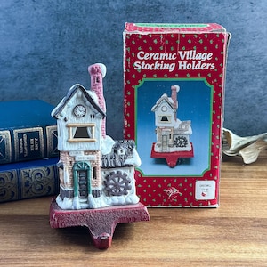 May include: A ceramic stocking holder shaped like a Grist Mill building with snow-dusted roof, a brick chimney, a water wheel, and a clock face. The item is displayed next to its red cardboard packaging box labeled Ceramic Village Stocking Holders.
