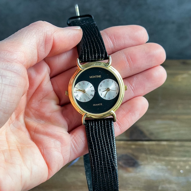 May include: A Montine quartz wristwatch featuring a gold-tone round case and a black dial with two silver sub-dials. The left sub-dial displays Roman numerals, while the right sub-dial shows Arabic numerals. It has a black textured leather strap.