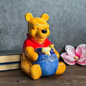 May include: A ceramic Winnie the Pooh figurine, featuring a yellow bear wearing a red shirt, holding a blue honey pot with the word "HUNNY". A small bee figurine sits on the pot. The figurine is on a wooden surface with books and a pink flower in the background.