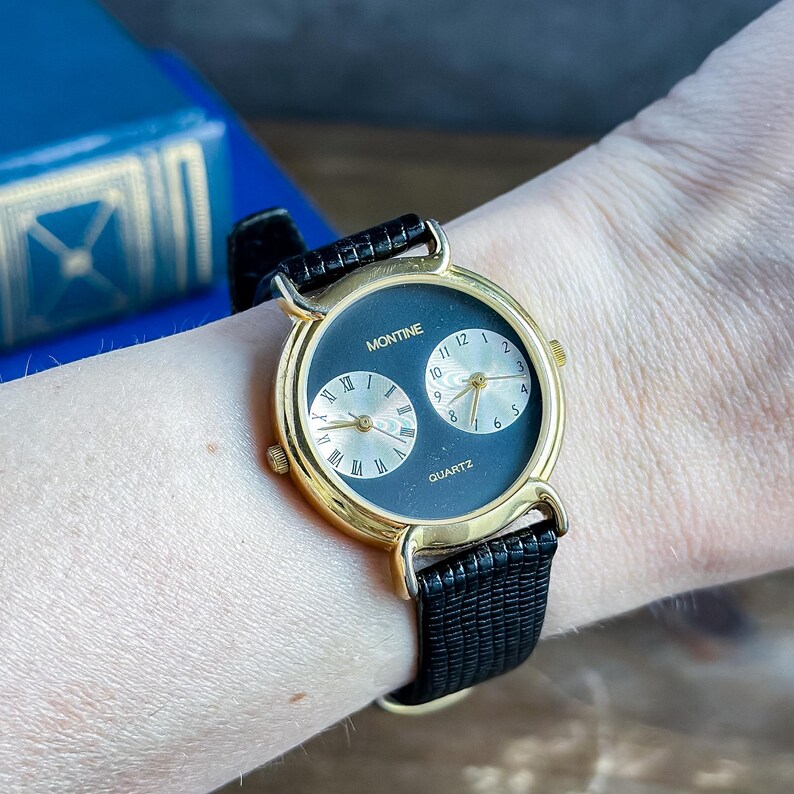 May include: A Montine quartz wristwatch featuring a gold-tone round case and a black dial with two sub-dials. One sub-dial displays Roman numerals, while the other shows Arabic numerals. The watch is fitted with a black textured leather strap.