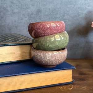 May include: A stack of three small, ceramic bowls in shades of pink, green, and tan. The bowls have a crackle glaze finish and are stacked on top of a blue book with gold-edged pages. The background is a muted gray.