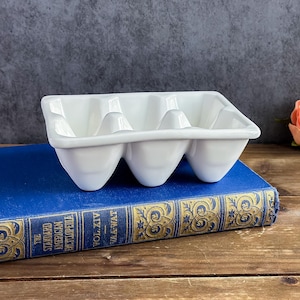 May include: A white ceramic egg holder with six compartments. The egg holder is rectangular with rounded egg-shaped indentations. It sits on a blue book with gold lettering. The book is titled "The Standard American Encyclopedia" and "Vol. XIV Vaa-Wat".