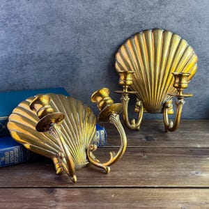 May include: Two brass seashell-shaped wall sconces with candle holders. The sconces have a detailed, ridged design and are mounted on a wooden surface. The candle holders are designed to hold two candles each, adding a touch of elegance to any space.
