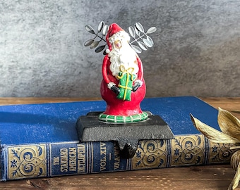 May include: A Christmas stocking holder featuring a Santa Claus figurine. The Santa is dressed in a red suit with a white beard and holds a green gift. The holder is black and sits on a blue book.