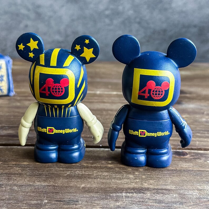 May include: Two blue Mickey Mouse figurines with yellow and red accents. The figurines have a television screen design on their heads with the number 40 and a Mickey Mouse head logo. The text "Walt Disney World" is printed on the base.