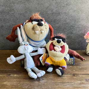 Peut inclure: Trois peluches représentant des personnages de Looney Tunes. Bugs Bunny est gris et blanc avec de longues oreilles. Taz, le Diable de Tasmanie, est marron et beige avec une tête noire. L'autre jouet Taz est dans une tenue jaune et violette.