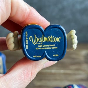 May include: A close-up of the base of a Vinylmation figure. The figure is dark blue with gold lettering that reads "Vinylmation" and "Walt Disney World 40th Anniversary Series". The figure has tan hands.