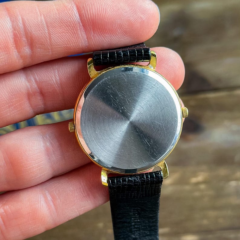 May include: A round wristwatch with a gold-tone metal case and a smooth, circular stainless steel back. The watch features a black textured leather strap with visible stitching and a small gold-tone crown on the side of the watch casing.