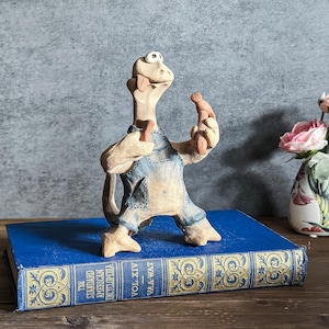 May include: A whimsical, hand-carved figurine of a creature with a long neck and arms, wearing denim overalls. The figurine is standing on a blue book with gold lettering. The creature is holding a number 7 in one hand and a small object in the other.