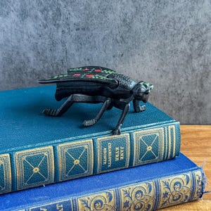 May include: A black cast iron insect figurine with green and red floral patterns on its wings, resting on a teal book titled Jane Eyre by Charlotte Bronte, which is stacked atop a blue book with gold decorative filigree patterns on the spine.