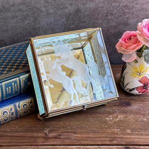May include: A square glass box with a gold-colored frame. The box features an etched design of two unicorns. The box is sitting on a wooden surface next to a vase of pink flowers and a stack of books.