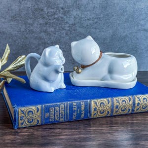 May include: Two white ceramic cat figurines. One is a small pitcher, the other is a planter with a brown collar and bells. Both are on a blue book with gold lettering. The background is a muted gray.