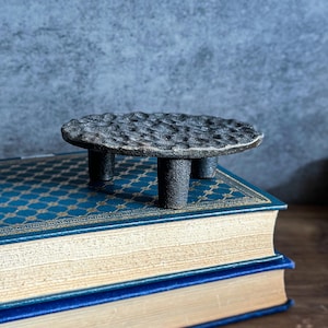 May include: A small, dark gray, textured metal table with three cylindrical legs. The tabletop has a hammered appearance. The table is displayed on top of a blue book with gold accents and a dark blue cover.