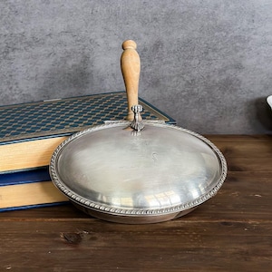May include: A silver-colored, oval-shaped serving dish with a wooden handle. The dish has a decorative rope-like border and a hinged lid. The handle is light brown. The dish is resting on a wooden surface.