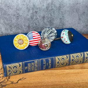 May include: A collection of decorative cabinet knobs with various designs, including a sunflower, an American flag, a globe, a ribbed glass knob, and a floral pattern. The knobs are displayed on a blue book with gold lettering.