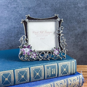 May include: A square metal picture frame with a dark finish and silver-toned scrollwork accents. The frame features purple and green enameled flower details at the corners. The insert reads Watch Hill Framers, genuine czech crystal, 3 x 3.