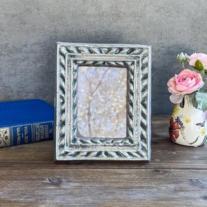 May include: A rectangular silver-toned picture frame with a textured, rope-like border design. The frame features a central opening with a light-colored, mottled insert. The frame is positioned next to a blue book and a floral vase with pink flowers.