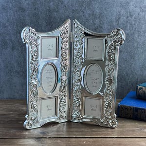 May include: A silver-toned metal hinged collage photo frame featuring ornate scrollwork and floral relief patterns. Each side contains three openings: two square 1.5 x 1.5 inch slots and one central oval 1.5 x 2 inch slot, all labeled with text.