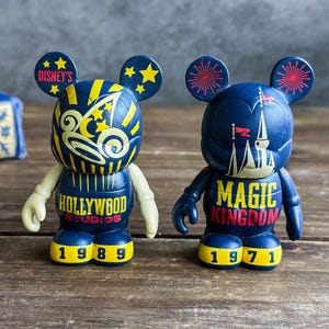 May include: Two navy blue Mickey Mouse figurines. One reads "Hollywood Studios 1989" with yellow and white accents. The other reads "Magic Kingdom 1971" with a castle design and red accents. Both have yellow feet and ears.