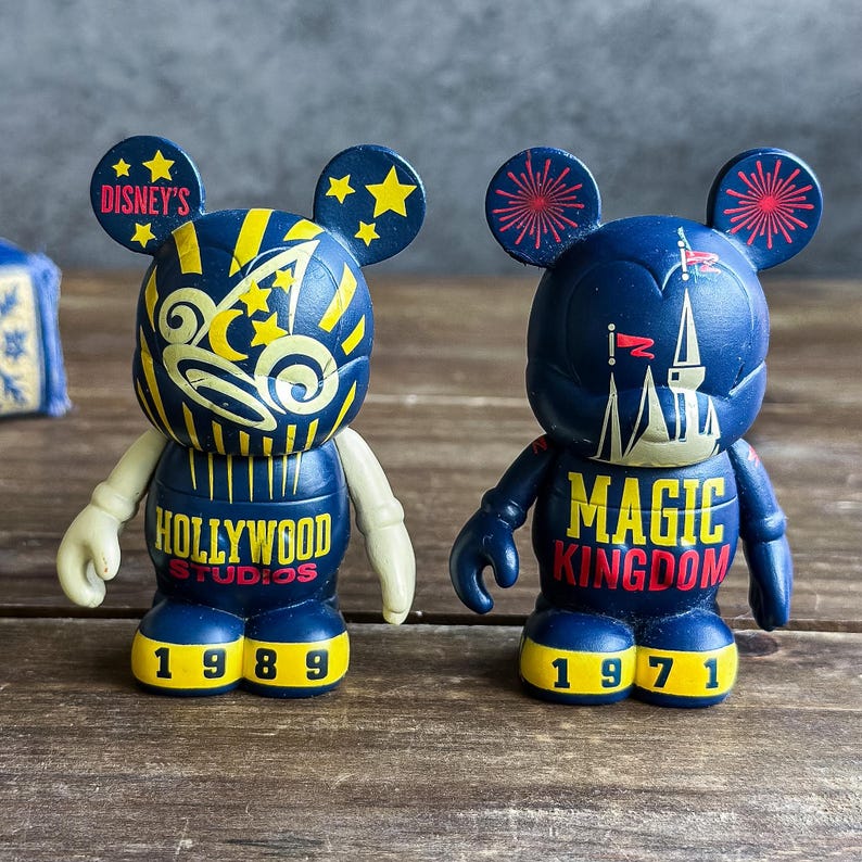 May include: Two navy blue Mickey Mouse figurines. One reads "Hollywood Studios 1989" with yellow and white accents. The other reads "Magic Kingdom 1971" with a castle design and red accents. Both have yellow feet and ears.