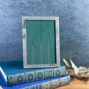 May include: A rectangular picture frame with a metallic silver finish featuring a marbled or brushed texture. The frame contains a green insert with a visible wood grain pattern. The frame is positioned vertically on top of two stacked books.