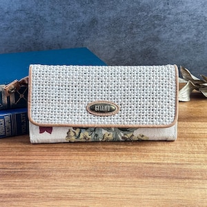 May include: A rectangular wallet featuring a light gray woven textured flap with a tan leather trim and a central oval metal plaque engraved with the word GITANO. The lower section displays a floral pattern in muted tones of yellow and green.