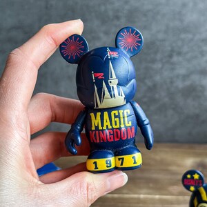 May include: A dark blue Mickey Mouse figurine with red and yellow details. The figurine displays the words "MAGIC KINGDOM" in yellow and red, and the number "1971" in yellow. The ears are decorated with red firework designs.