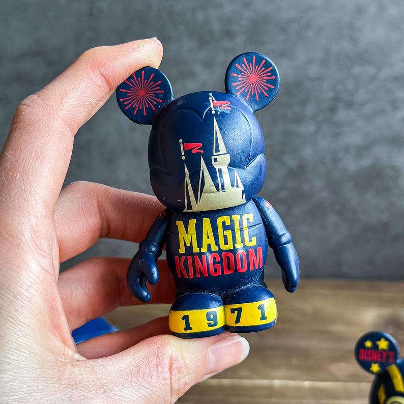 May include: A dark blue Mickey Mouse figurine with red and yellow details. The figurine displays the words "MAGIC KINGDOM" in yellow and red, and the number "1971" in yellow. The ears are decorated with red firework designs.