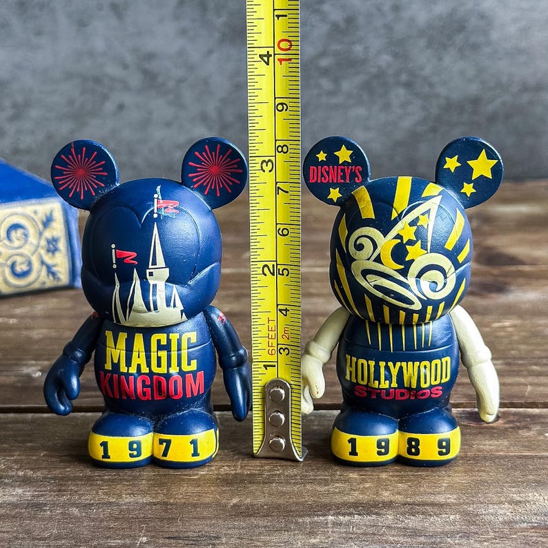 May include: Two Disney-themed figurines, each approximately 10 cm tall, with Mickey Mouse ears. One is navy blue with "MAGIC KINGDOM 1971" text, the other is navy blue and yellow with "HOLLYWOOD STUDIOS 1989" text. A yellow ruler is placed next to the figurines.