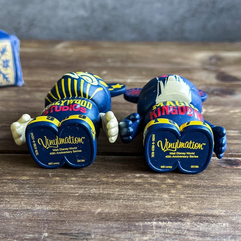 May include: Two blue Vinylmation figures from the Walt Disney World 40th Anniversary Series. One figure has "Hollywood Studios" printed on it, and the other has "Magic Kingdom". Both figures have white hands and yellow accents.