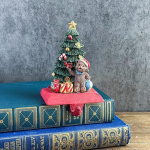 May include: A cast iron Christmas stocking holder featuring a green pine tree decorated with gold ornaments, red and white candy canes, and a gold star topper. A brown teddy bear wearing a red Santa hat and blue scarf stands beside wrapped gifts.