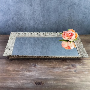 May include: A rectangular mirrored tray featuring an ornate, metallic silver-toned frame with intricate scrollwork patterns along the edges. A single, large, soft peach-colored peony flower rests on the surface, reflecting in the mirror.