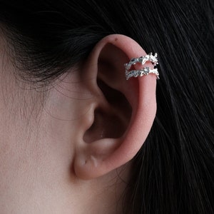 May include: A silver ear cuff with a delicate, intricate design. The cuff is made of two overlapping bands of metal, each with a unique pattern. The ear cuff is a stylish and modern accessory that can be worn on its own or with other earrings.