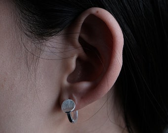 May include: Close-up of a silver earring on an ear. The earring has a small, textured, circular design attached to a hoop. The earring is worn on the ear of a person with dark hair.