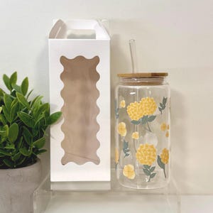May include: A clear glass tumbler with a bamboo lid and straw, decorated with yellow floral designs and green leaves. A white box with a wavy window is next to the tumbler. A small potted plant is in the background.