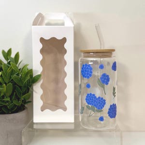 May include: A clear glass tumbler with a bamboo lid and a clear straw. The tumbler is decorated with blue hydrangea flowers and green leaves. A white box with a scalloped window is to the left of the tumbler. A small potted plant is in the background.