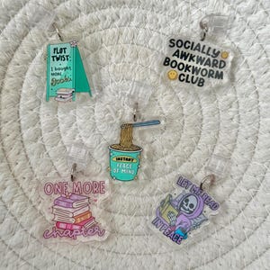May include: A selection of clear acrylic badge reels featuring book-related designs. The designs include phrases such as "Plot Twist," "Socially Awkward Bookworm Club," and "One More Chapter," alongside illustrations of books and a reading skeleton.