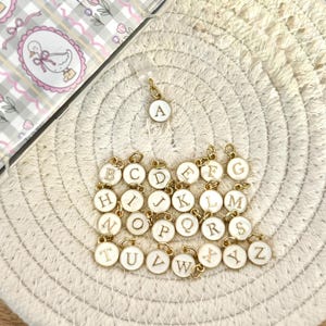 May include: A selection of gold-tone alphabet charms with white enamel centres. Each charm displays a different letter from A to Z. The charms are arranged on a cream-coloured woven surface, with a decorative case in the background.