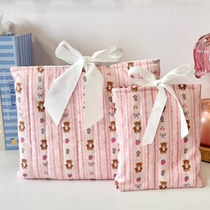Puede incluir: Dos bolsas de tela rosa con lazos de cinta blancos. Las bolsas presentan un patrón repetido de osos de peluche marrones, lazos y fresas sobre un fondo a rayas. Las bolsas son de diferentes tamaños.