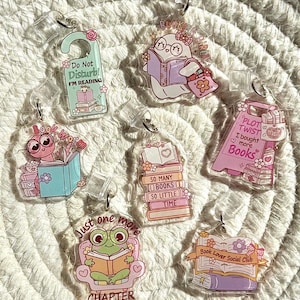 May include: A selection of clear acrylic keyrings with book-related designs. Each keyring showcases a unique illustration, such as a frog reading, a ghost reading, and stacks of books. Text includes "Do Not Disturb! I'm Reading" and "Book Lover Social Club".